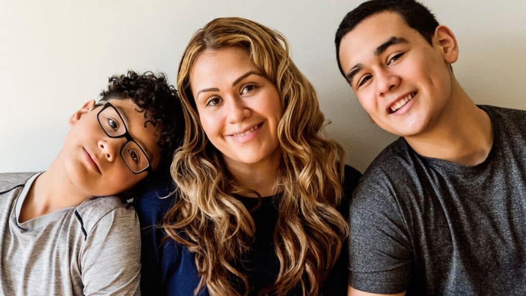 A smiling mom sits between her two sons. One boy with glasses leans his head on her shoulder, and the other boy smiles at the camera.