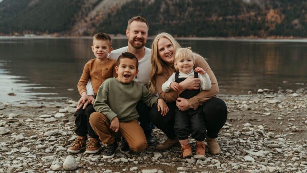 A family of five sitting on a rocky beach with a lake and mountains in the background.