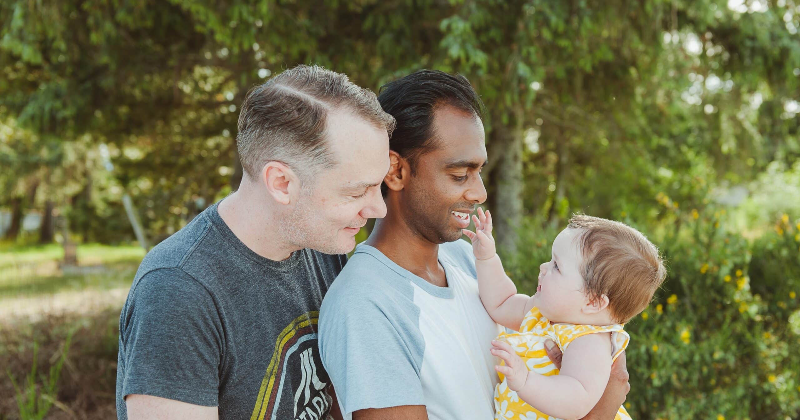 A same-sex couple smiling and holding their baby girl in their arms.