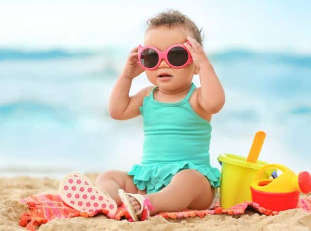 A baby in a turquoise swimsuit and pink sunglasses sits on the beach with a towel and sand toys.