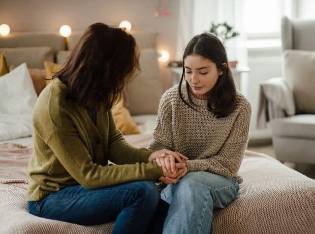 A mom sits on a bed, holding hands with her teenage daughter.
