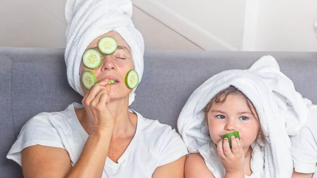 A mother and daughter wearing towels on their heads and cucumber slices on their faces, enjoying a self-care moment together.