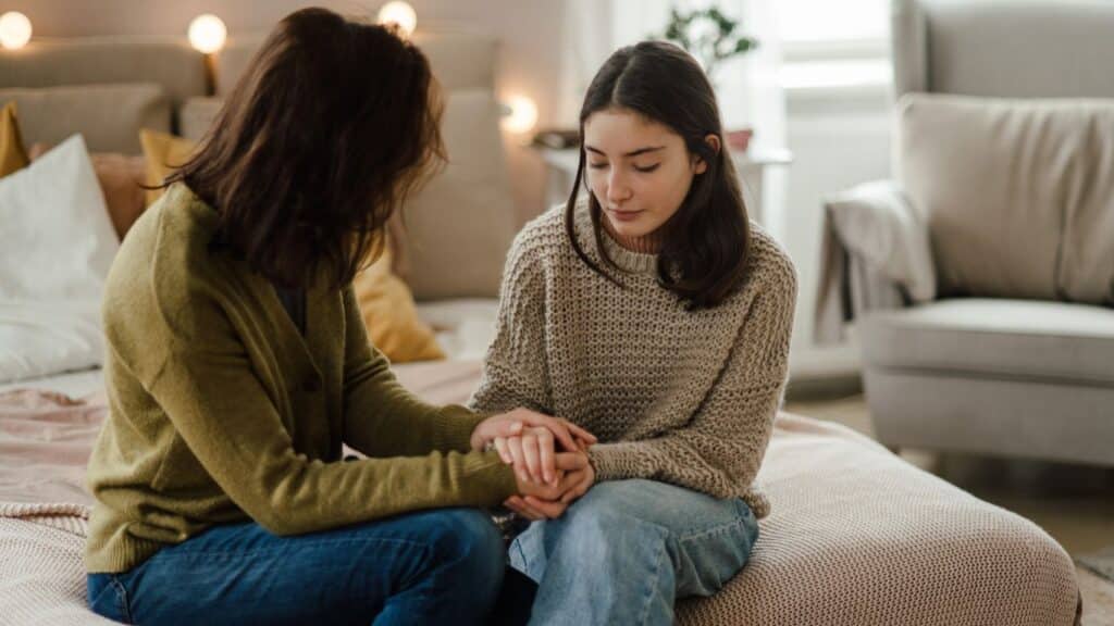 A parent sits with a teenage girl on a bed, holding her hands in a supportive, caring way.