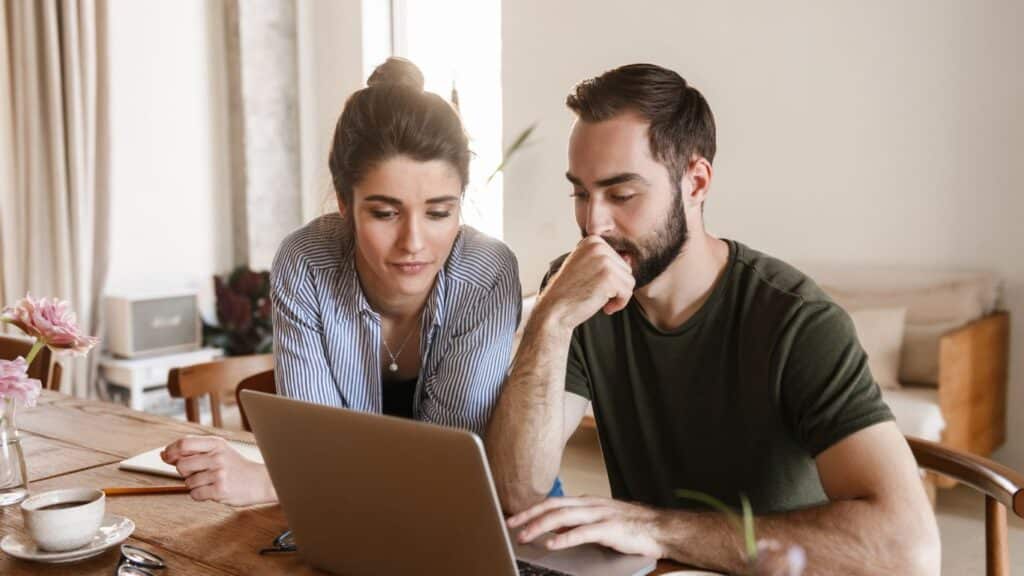 Two adults sit side by side at a table, looking thoughtfully at a laptop.