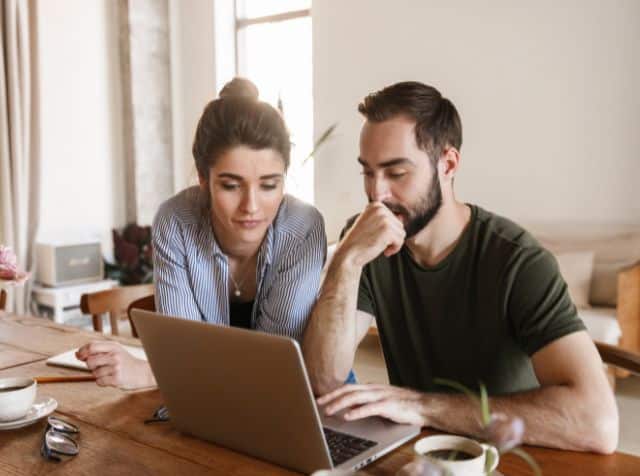 A couple sit side by side at a table, looking thoughtfully at a laptop.